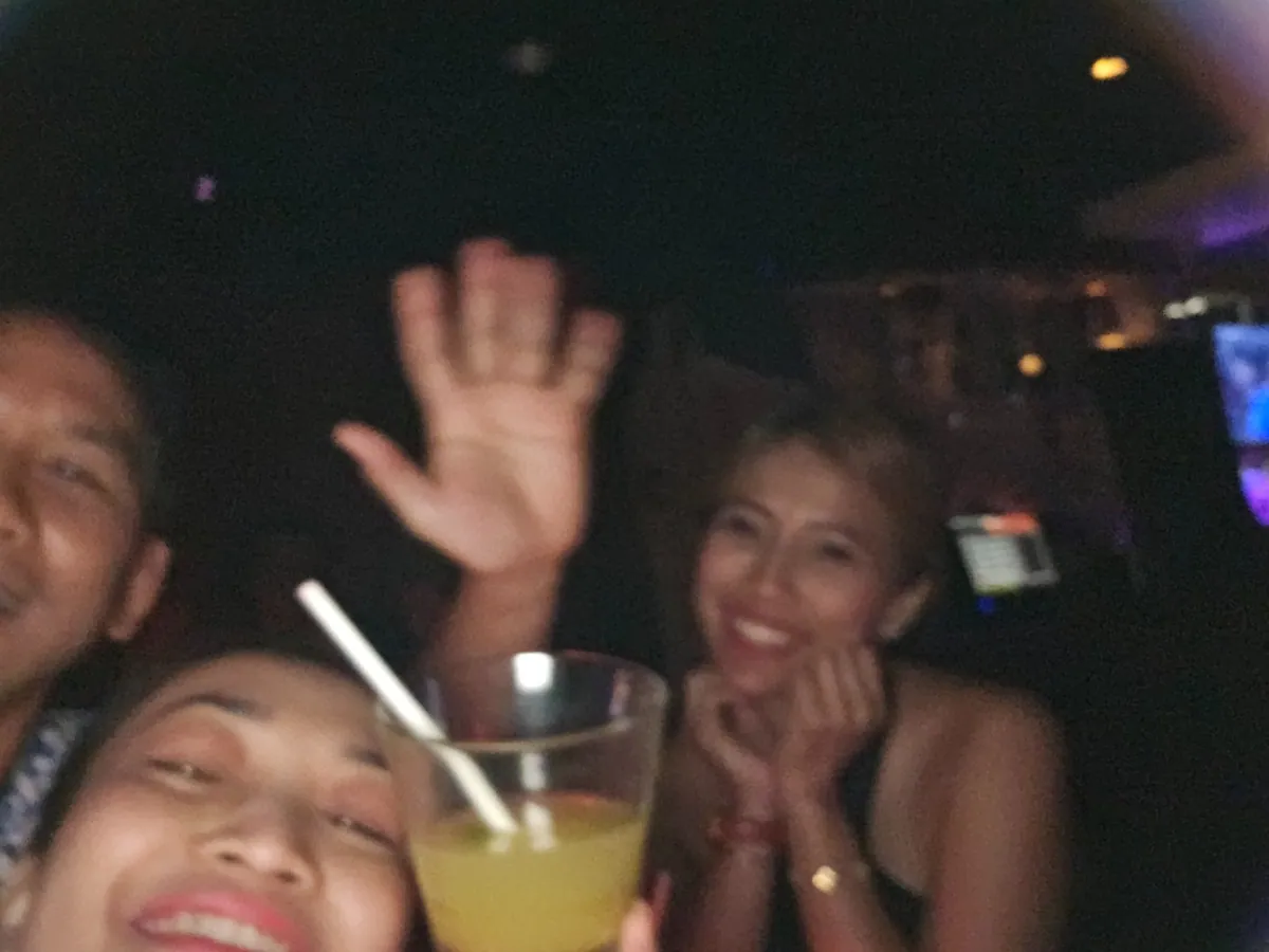 Three guests at a dark bar table, one waving at the camera while another holds a cocktail.