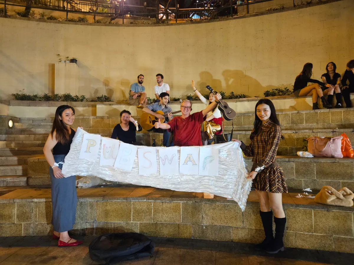 A group on stone amphitheatre steps holding a hand-painted PubSwap banner while a guitarist plays behind them.