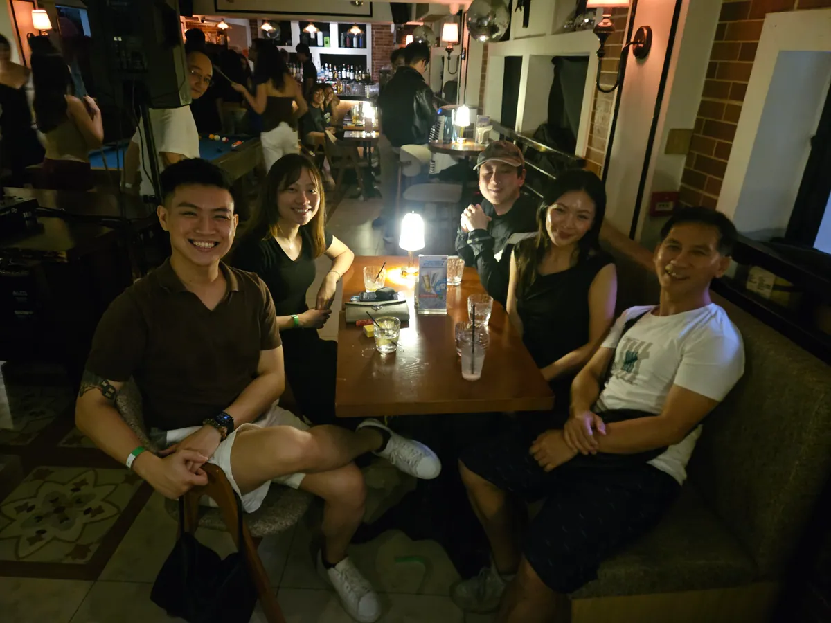 Five guests around a wooden table in a dim bar, posing with cocktails and a lit candle.