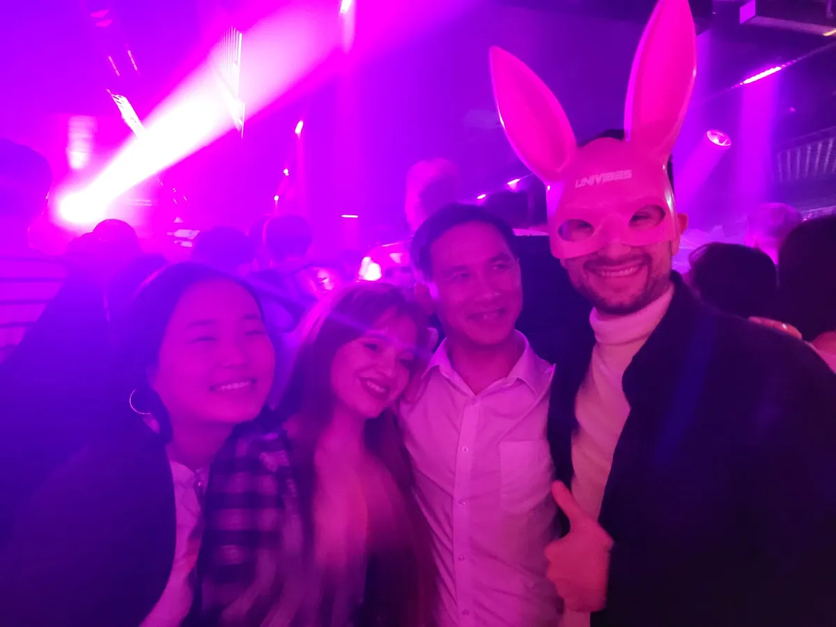 Four guests in a purple-lit club, one wearing a pink bunny-ear headband and giving a thumbs up.