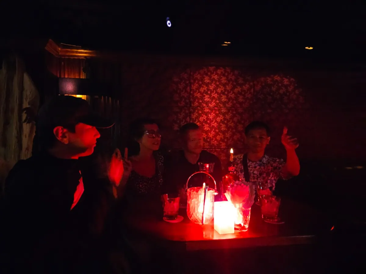 Four guests around a table in a red-lit bar, with a lantern and candles between their drinks.