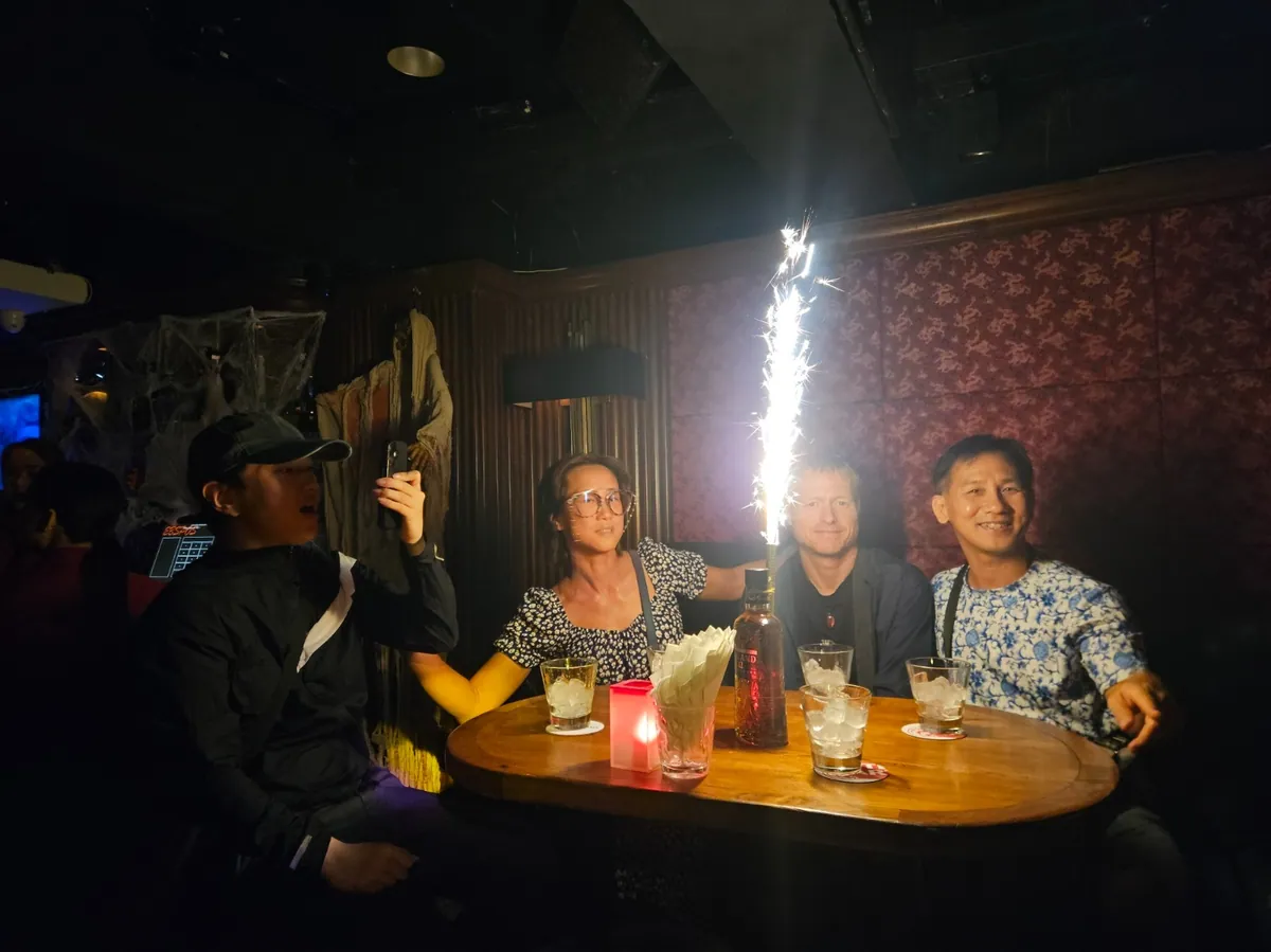 Four guests at a round wooden table in front of red patterned wallpaper while a sparkler burns on a bottle.