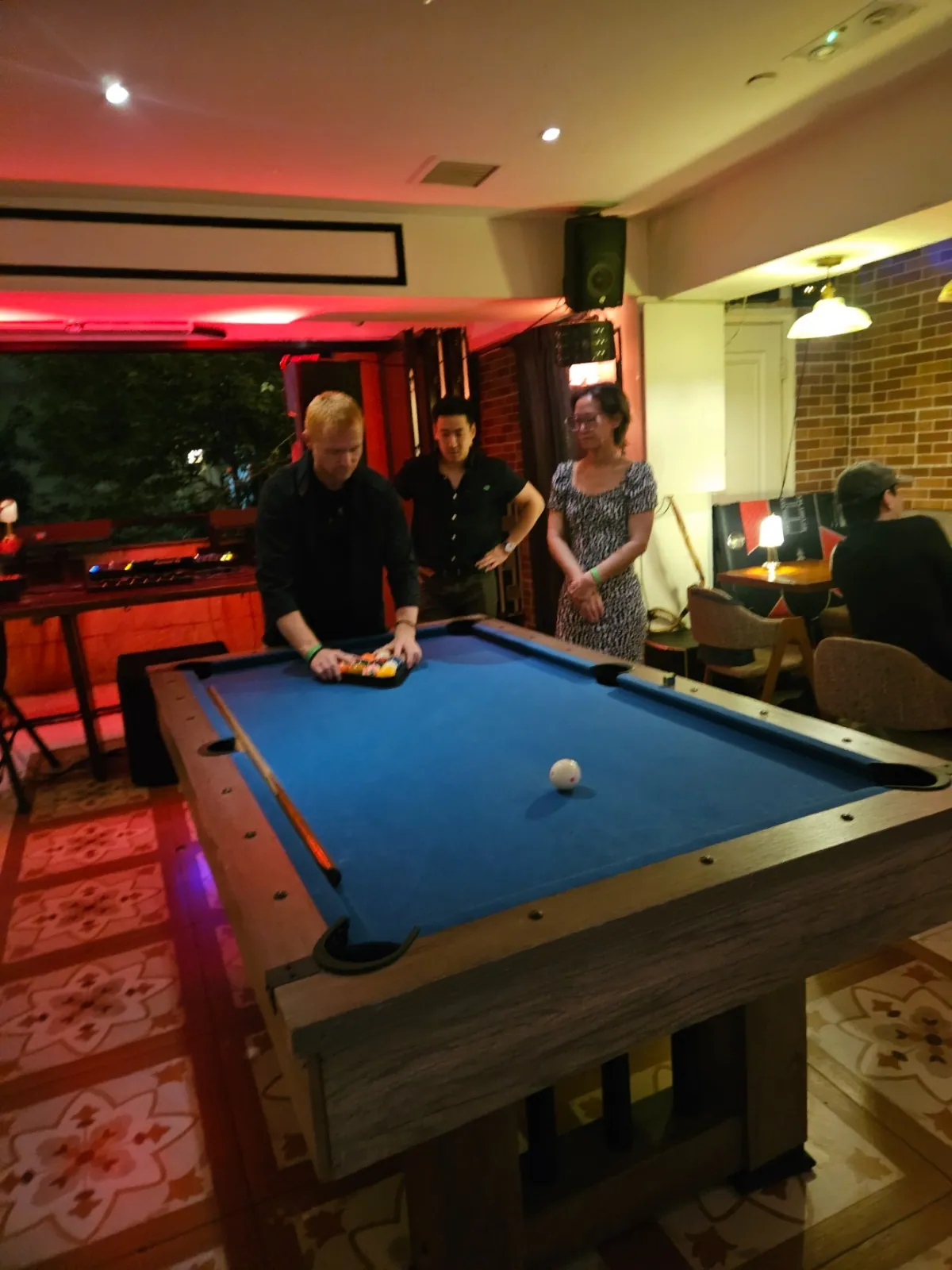 A guest racking pool balls on a blue table while two others watch in a bar lit with red neon.