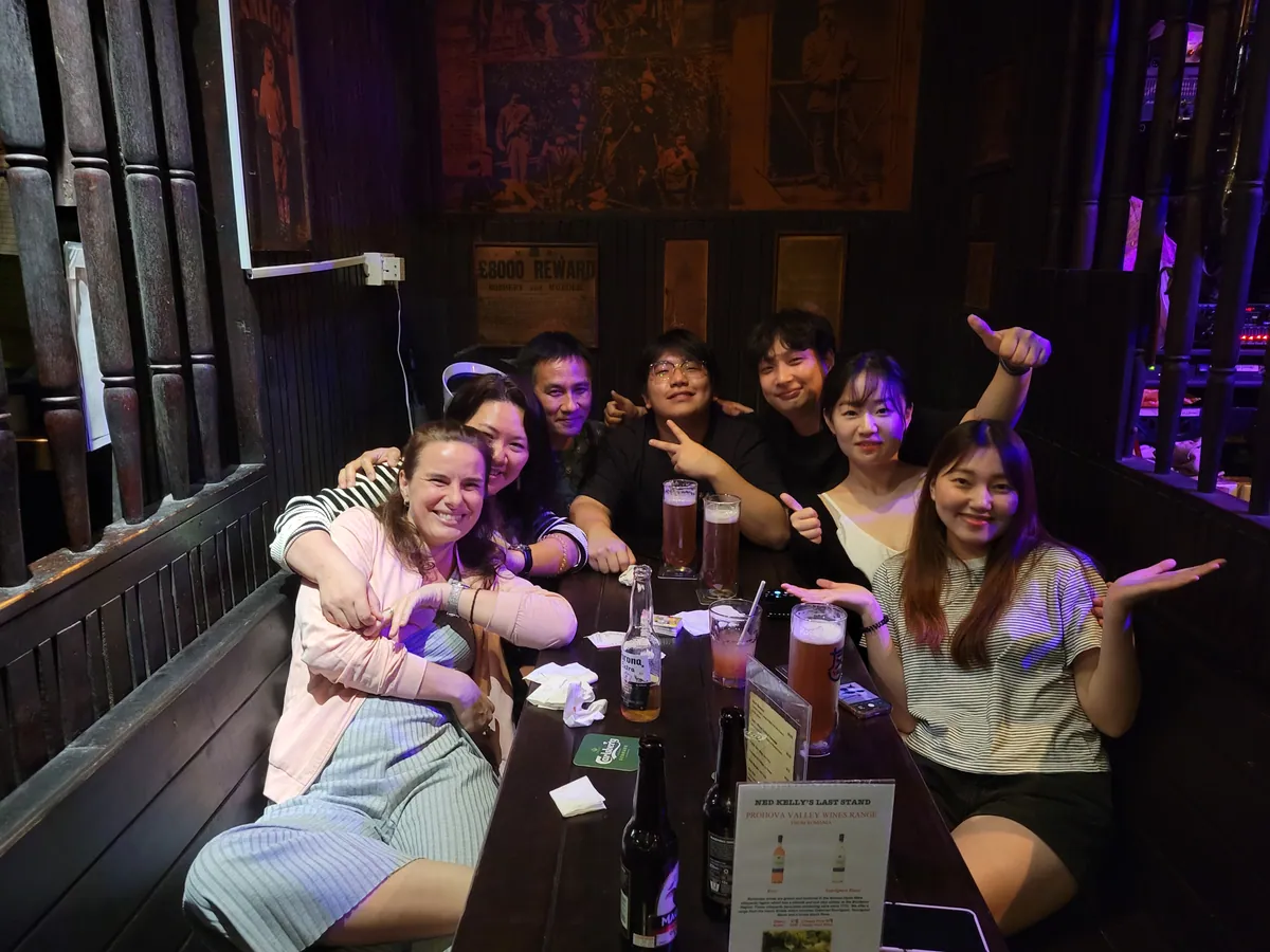 Eight guests crammed into a wooden pub booth with their arms around each other, pints and cocktails lined up on the table.