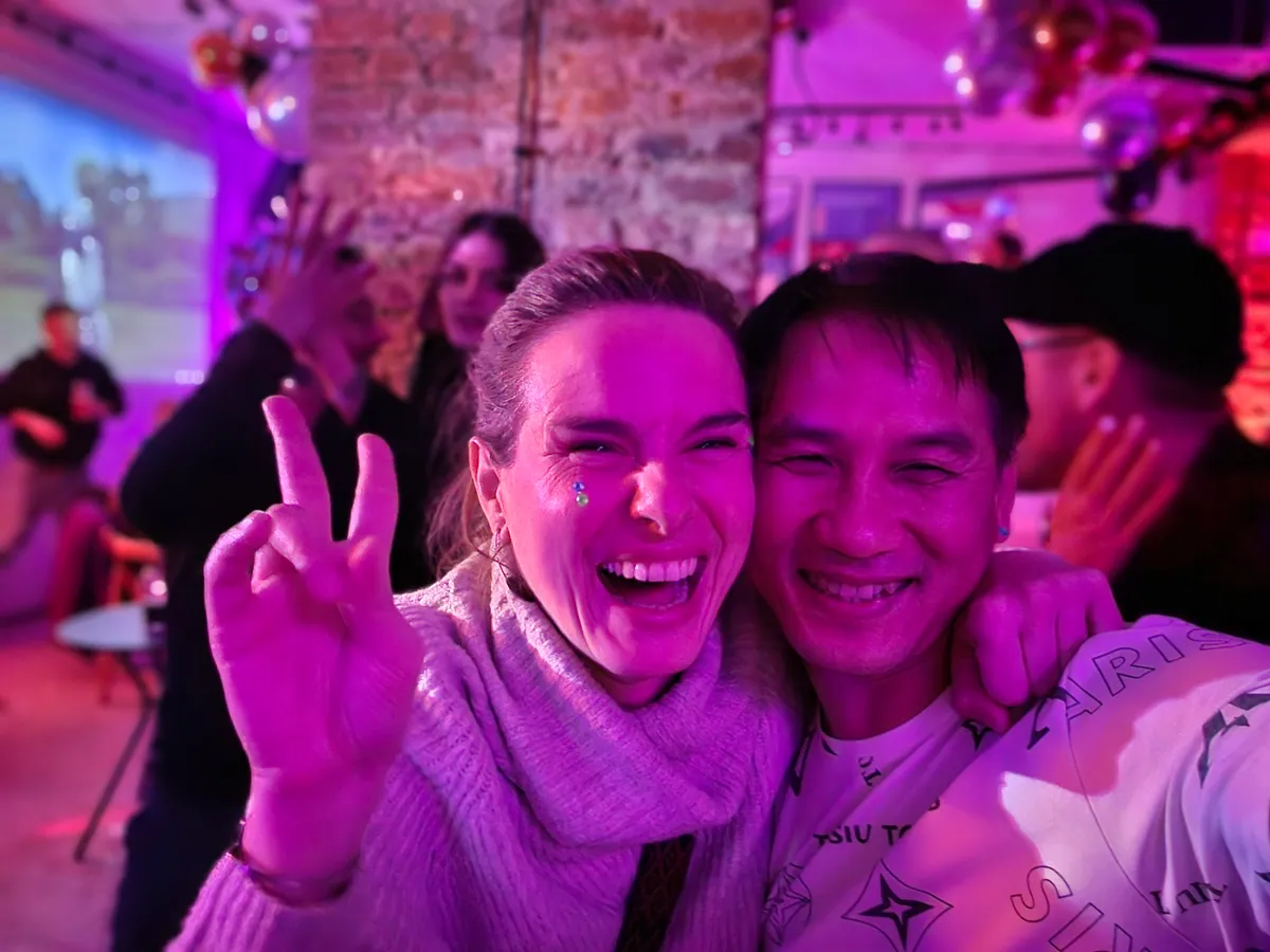 Two guests grinning under magenta club lights in front of a brick wall, one throwing a peace sign.