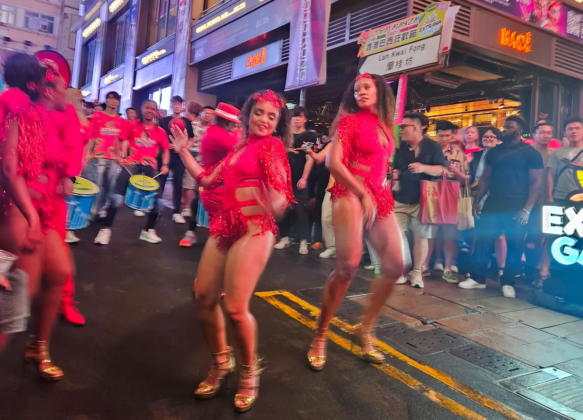 Two dancers in red fringed costumes perform in a Lan Kwai Fong street while drummers in red shirts and a crowd look on.